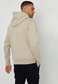 Sudadera beige con capucha ajustable, puños y dobladillo acanalados. Tejido suave, diseño liso y corte relajado, llevada sobre pantalones cargo oscuros.