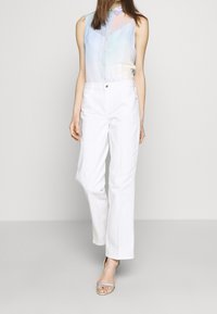 Blusa trasparente senza maniche in colori pastello con bottini frontali e un colletto sottile, abbinata a pantaloni bianchi a gamba dritta e sandali argento.