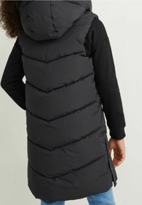 Czarna puchowa kamizelka z kapturem, z charakterystycznym przeszyciem w zygzak, długim krojem, bocznymi rozcięciami oraz gładkim, błyszczącym wykończeniem. Noszona na czarnej koszuli z długim rękawem.