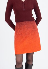 Femme portant un haut long à manches ribbed bourgogne rentré dans une mini-jupe orange texturée, avec des collants noirs et des bottes noires.
