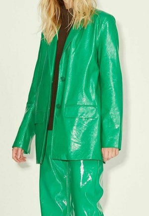 Kobieta w błyszczącym zielonym skórzanym żakiecie i dopasowanych spodniach na ciemnobrązowym topie, stojąca na jednolitym jasnym tle.