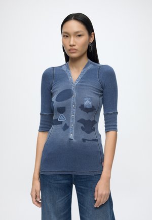 Donna con lunghi capelli neri indossa una camicia blu aderente con bottoni, motivi astratti e maniche a lunghezza media, abbinata a jeans blu.