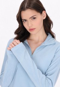 Mujer joven con pelo castaño que lleva puesto un pulóver de felpa azul claro, con las manos juntas cerca del pecho, mirando hacia adelante con una expresión neutral.