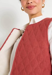 Gilet matelassé de couleur rouille avec une texture ondulée, doublé d'un matériau crémeux doux. Il présente un col rond et est porté sur une chemise blanche.