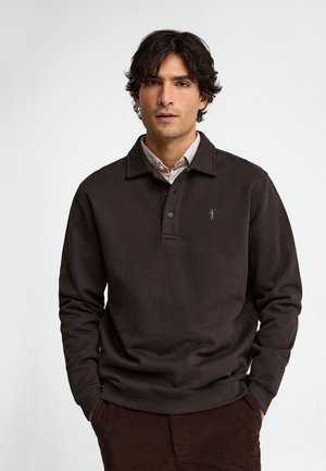 Joven con cabello oscuro que lleva un jersey marrón oscuro sobre una camisa clara, de pie con las manos en los bolsillos frente a un fondo liso.