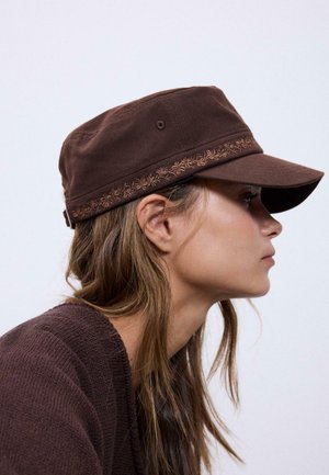 Giovane donna con capelli lunghi, indossa un berretto marrone ricamato e un maglione marrone coordinato, guarda verso destra su uno sfondo chiaro e uniforme.