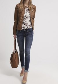 Brun läderjacka, mönstrad blommig blus, blå skinny jeans, beige klackar och en brun handväska med kedjeaccent. Avslappnad outfit.