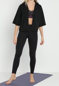 Černá zkrácená mikina s širokými rukávy, předním zipem a texturovanou tkaninou. Kombinováno s přiléhavými černými legínami s akcenty z síťoviny.