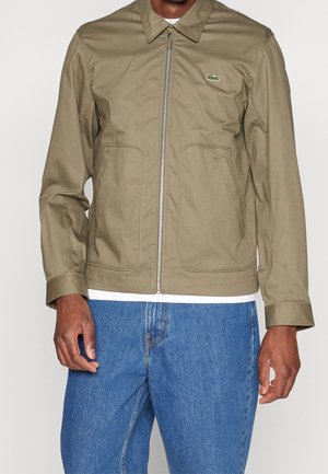 Man in een olijfgroene jas met rits en kraag, met twee voorzakken, gecombineerd met een blauwe spijkerbroek en een wit ondershirt.