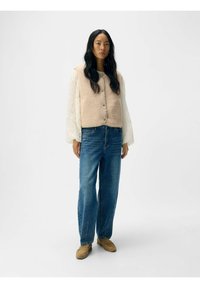 Beige fleece vest med knapper, båret over en hvid mønstret bluse; blå lige ben jeans; brune sko. Enkle, teksturerede stofdetaljer.
