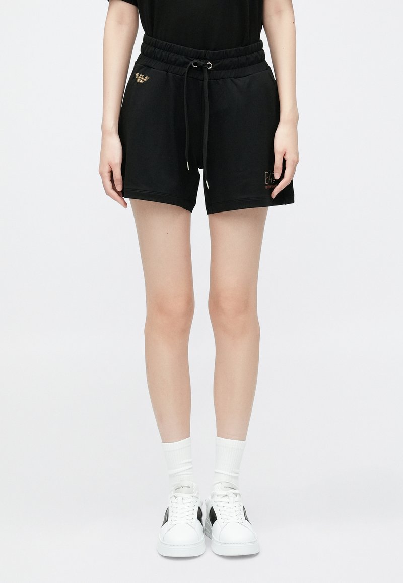 Fekete rövidnadrág elasztikus derékpánttal és húzózsinórral, arany logó részletekkel. Fehér sportcipővel és bokazoknival párosítva.