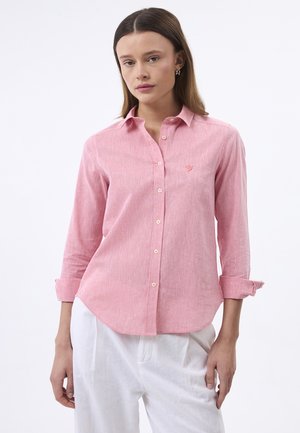 Femme portant une chemise rose à manches longues boutonnée avec des poignets retroussés et un pantalon blanc sur un fond uni.