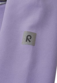 Tissu violet avec une texture lisse, arborant un logo rectangulaire argenté avec un 'R' intégré dans le coin. Vue en gros plan.