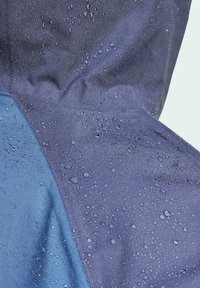 Chaqueta resistente al agua en azul marino y azul con gotas de agua visibles sobre una tela texturizada, mostrando una superficie suave y duradera.