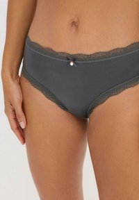 Donna che indossa un perizoma di pizzo grigio scuro con un piccolo fiocco e un dettaglio a charm in vita.