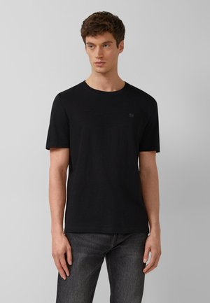 Młody mężczyzna z krótkimi brązowymi włosami, ubrany w jednolitą czarną koszulkę z krótkim rękawem i ciemnoszare dżinsy, stojący na jasnoszarym tle.