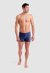 Herren Badeshorts in Marineblau aus glattem Stoff, mit einem weißen Logoaufdruck an der Seite. Für eine enganliegende Passform und eine mittlere Oberschenkellänge gestaltet.