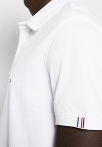 Homme portant un polo blanc à manches courtes avec un col et de petites rayures rouges, blanches et bleues sur le bord des manches.