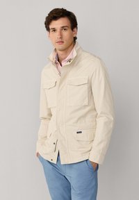 Jonge man met een beige jas met klepzakken, een roze overhemd en blauwe broek, staand tegen een neutrale grijze achtergrond.