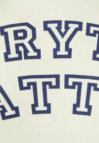 Bílá tkanina s tmavě modrým textem ve tučných, blokových písmenech, který říká "RYT ATT." Textura vypadá jako pletená s mírně vyvýšeným vzorem.