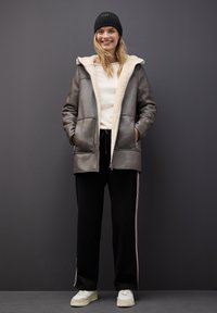 Giubbotto in finta pelle marrone con un rivestimento in finta pelliccia color crema, due tasche frontali, abbinato a pantaloni sportivi neri con righe laterali e sneakers bianche.