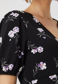 Svart klädestyg med ett blommönster av vita och lila blommor. Plagget har en V-ringning och puffärmar.