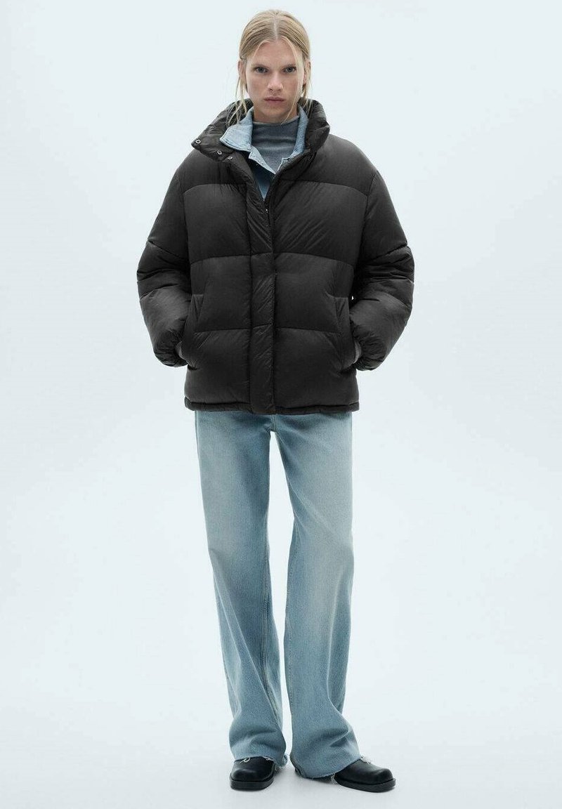 Sort pufferjakke med høj krave, quiltet design og lynlås. Parret med lyseblå vide jeans og sorte sko.