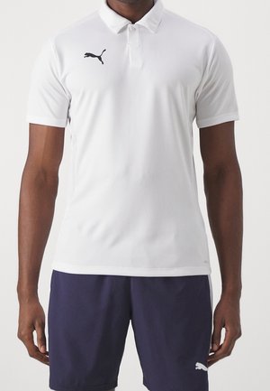 Mann trägt ein weißes Puma-Poloshirt und dunkelblaue Puma-Shorts und steht vor einem einfarbigen Hintergrund.