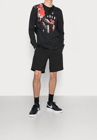 Mies, joka pukeutuu mustaan pitkähihaiseen paitaan, jossa on pikselöity punainen graafinen kuvasto, mustiin shortseihin, mustiin Nike-tennareihin, mustiin sukkiin ja mustaan olka-/rintalaukkuun.