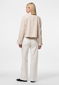 Una donna con i capelli biondi lisci indossa una giacca beige, jeans bianchi a gamba larga e scarpe nere, in piedi con le spalle rivolte verso la macchina fotografica.