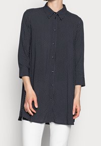 Navy blue button-up shirt with white polka dots, long sleeves, and a classic collar; smooth fabric, slightly loose fit, mid-length design.