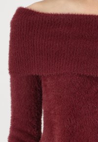 Bordo pulover s off-shoulder dizajnom s rebrastim izrezom i pahuljastom teksturom. Sadrži mekan i topao materijal, idealan za hladnije vrijeme.