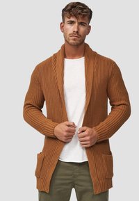Strikket cardigan i varm brun, med sjalskrave, ribbede manchetter og to forlommer. Den strukturerede stof giver dybde.