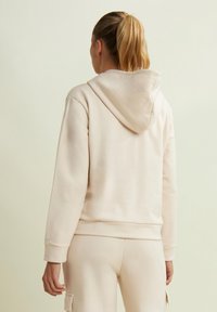 Femme avec une queue de cheval blonde portant un hoodie beige et un pantalon de jogging assorti, debout et faisant face à un fond uni.