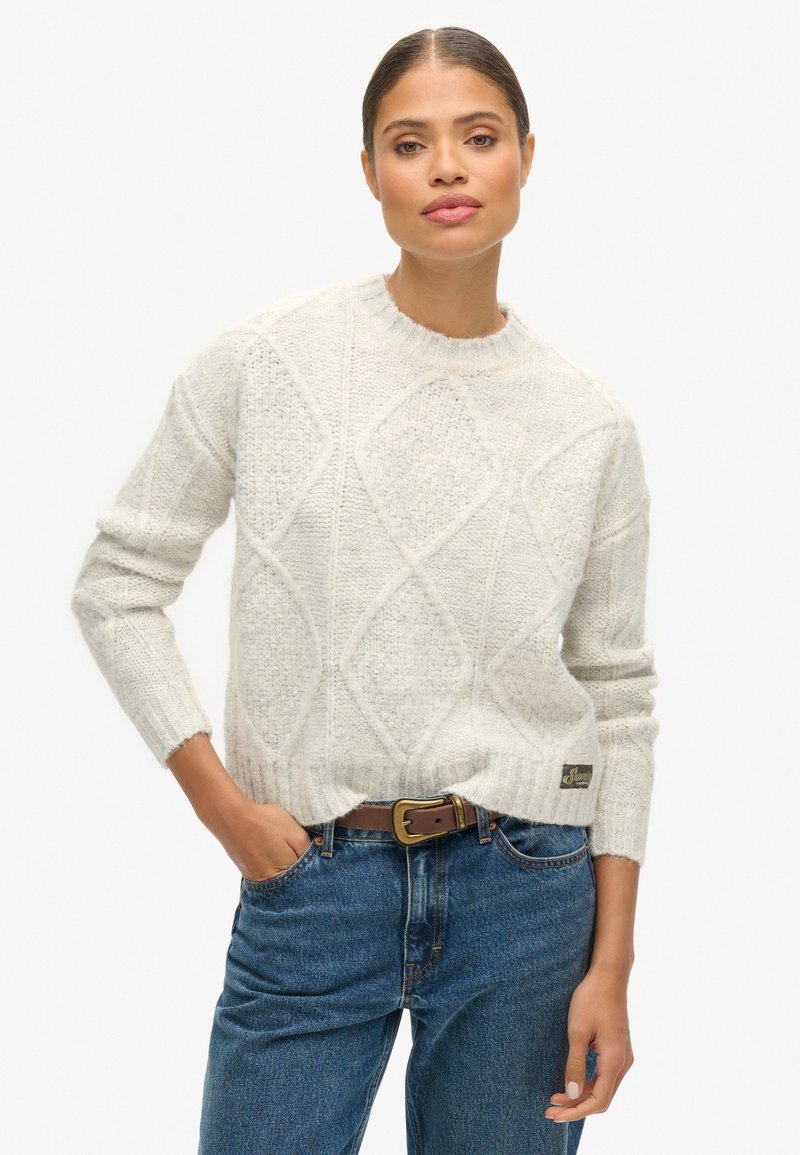 Pull crème avec un motif en losanges, tricot texturé et bords côtelés. Assorti à un jean bleu taille haute et une ceinture marron.
