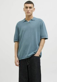 T-shirt à manches courtes et col en V en tissu bleu pastel avec une texture lisse ; associé à un pantalon en denim foncé, avec une coupe décontractée et des coutures visibles.