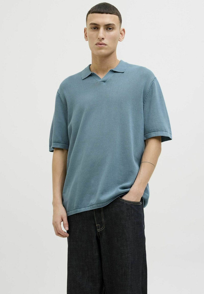 T-shirt à manches courtes et col en V en tissu bleu pastel avec une texture lisse ; associé à un pantalon en denim foncé, avec une coupe décontractée et des coutures visibles.