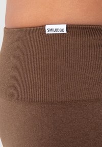 Leggings marroni a coste con una texture liscia. Presentano un'etichetta bianca con la scritta "SMILODOX" sulla vita.