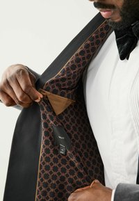 Schwarze Blazerjacke mit einem gemusterten Innenfutter, das ein geometrisches Design in Brauntönen aufweist, und einem sichtbaren Etikett mit Größendetails.