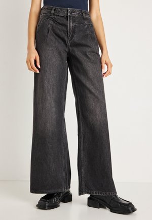Pantaloni a gamba larga neri in denim a vita alta con pieghe frontali, orlo grezzo e una texture morbida. Abbinati a scarpe nere con tacco a blocco.