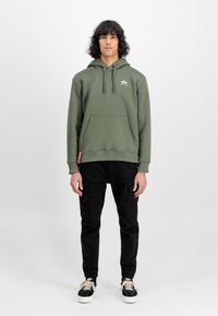 Olivgrüne Kapuzenjacke mit Kängurutasche und Kordelzügen. Schwarze Cargo-Hosen mit Seitentaschen. Schwarze und weiße Sneakers runden das Outfit ab.