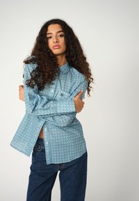 Light blue collared shirt with white circular patterns, button front, long sleeves, and a small chest pocket, paired with dark blue jeans.