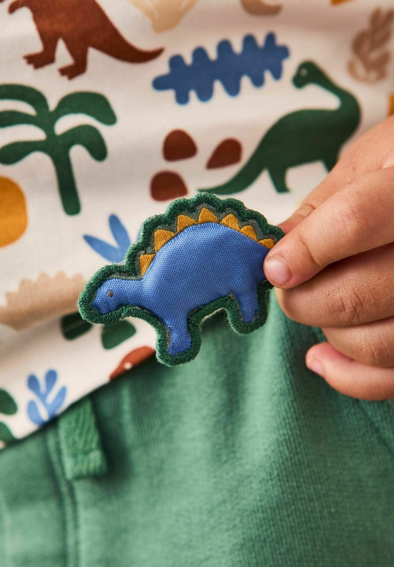 Blå stof dinosaurusmærkat med grøn kant og orange detaljer, holdt imod en mønstret skjorte med træer og dinosaurer, over grøn stof.