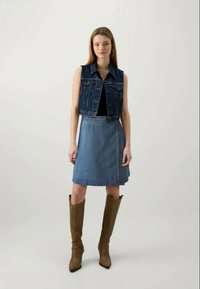 Colete de ganga com um design cropped, usado sobre uma blusa preta. Saia de ganga em forma de A, até ao joelho. Botas castanhas até ao joelho com uma forma ajustada.
