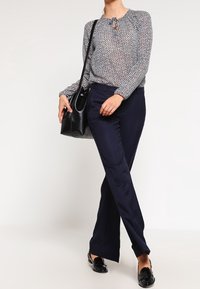 Top a maniche lunghe con un motivo geometrico, abbinato a pantaloni blu navy. Il modello tiene in mano una borsa nera e indossa mocassini neri lucidi.
