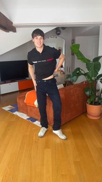 Νεαρός άνδρας που χαμογελά, φοράει μαύρο πουκάμισο Tommy Hilfiger και σκούρο τζιν, στέκεται στο σαλόνι κοντά σε πορτοκαλί καναπέ και μεγάλο γλαστρικό φυτό.