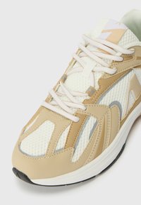 Zapatilla deportiva beige con parte superior de malla y acentos de cuero. Presenta cordones blancos, un cuello acolchado y una suela de goma texturizada.