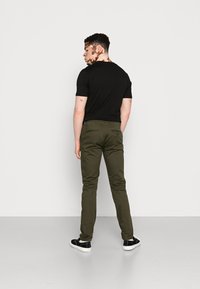 Camiseta negra de manga corta combinada con pantalones ajustados verde oliva y zapatillas negras, vista desde atrás sobre un fondo blanco.