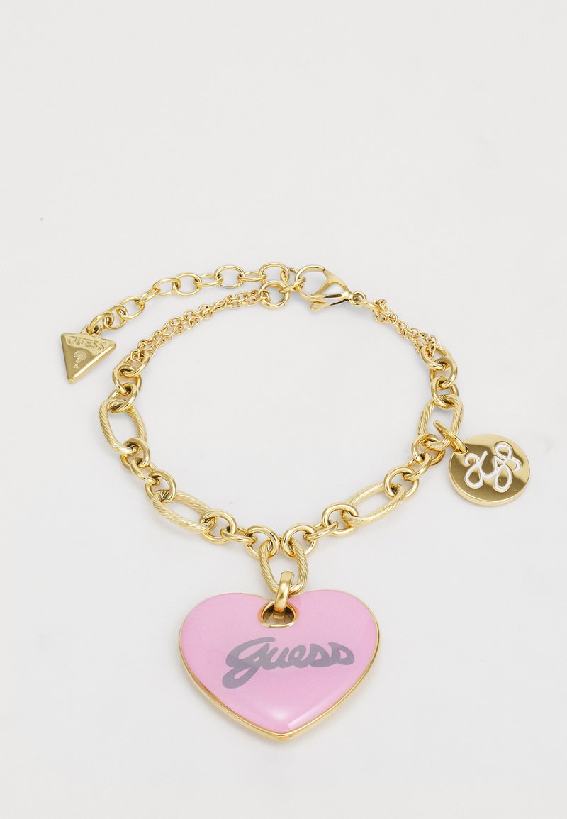 Brățară din aur cu un charm în formă de inimă roz, având logo-ul "Guess" în gri, și un al doilea charm rotund cu un emblema stilizată "G".