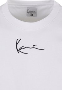 T-shirt blanc col rond avec logo « Kani » noir en écriture cursive brodé sur la poitrine et une étiquette taille large à l'intérieur de l'encolure.